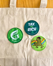 Three woven patches featuring three designs: a Green logo with leaves, a Tax the Rich slogan, and a Tuatara with a rainbow in the background. 