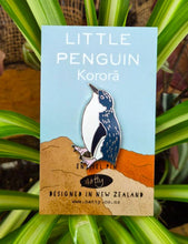 Pin with a kororā/little blue penguin design on a blue card, surrounded by green plants.